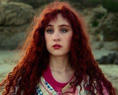 a woman with long red hair standing in front of rocks and looking at the camera