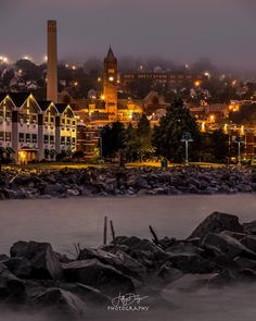 the city is lit up at night by the water's edge with rocks in front of it