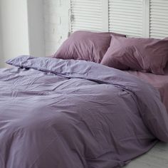 a bed with purple sheets and pillows in a white room next to a window that has shutters