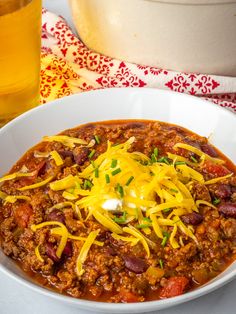 a white bowl filled with chili and cheese next to a glass of beer on a table