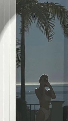 a man in a bathing suit standing next to a palm tree and looking out at the ocean