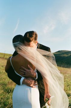 a bride and groom embracing each other on their wedding day in a field with mountains behind them