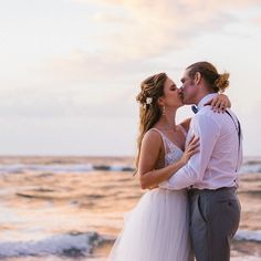 a bride and groom kissing on the beach at sunset with waves crashing in behind them