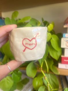 a person holding up a small cup with the word mum on it in front of some plants