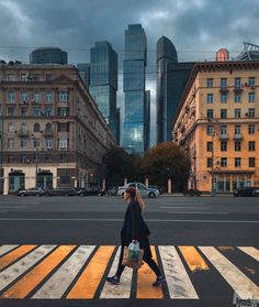 a woman walking across a cross walk in front of tall buildings on a cloudy day