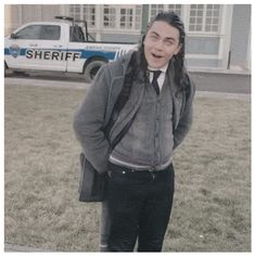 a man with long hair wearing a gray jacket and tie standing in front of a police car