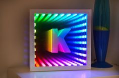 a lit up letter k in front of a blue vase on a white table top