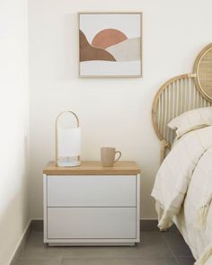 a white nightstand with a cup on it next to a bed and a painting hanging above it