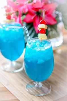 two glasses filled with blue liquid sitting on top of a wooden table next to flowers