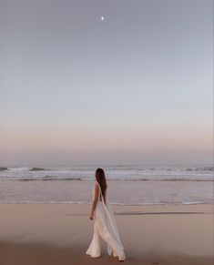 a woman in white dress walking on beach next to the ocean at dusk with full moon