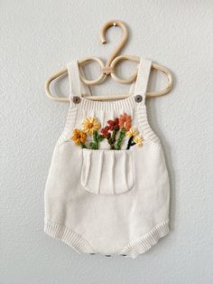 a white knitted romper with flowers in the pocket hanging on a wooden hanger