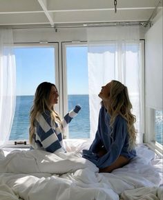 two women sitting on a bed in front of large windows looking out at the ocean