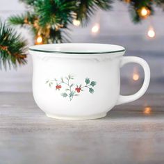 a white cup sitting on top of a wooden table next to a christmas tree with lights in the background