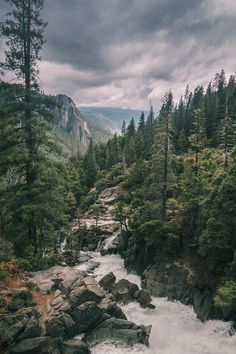 a river running through a forest filled with rocks and trees on a cloudy day in the mountains