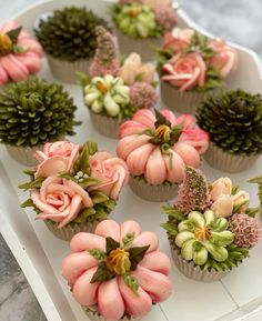 cupcakes decorated with pink and green flowers on a white tray next to pine cones