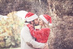 a man and woman in santa hats kissing under a tree with snow falling on them