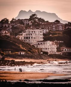 people are walking on the beach in front of some houses and water with mountains in the background