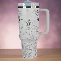 a white travel mug with mickey mouse drawings on the front and sides, sitting on a wooden table