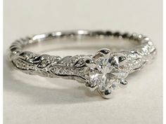 a white gold engagement ring with an intricate design on the band and a round diamond center stone