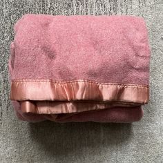 the pink blanket is folded on top of the gray carpeted floor and has a satin edge