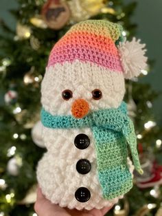 a hand holding a knitted snowman in front of a christmas tree