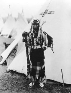 an old black and white photo of a native american man standing in front of a teepee