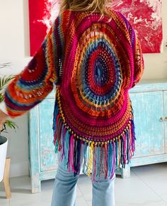 a woman standing in front of a painting wearing a colorful crocheted sweater with fringes