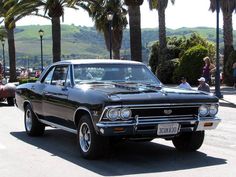 an old black car is driving down the street with palm trees in the back ground