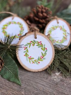 three wooden ornaments with holly and berries on them, sitting next to pinecone cones