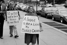 two women holding signs while walking down the street with other people on the sidewalk behind them