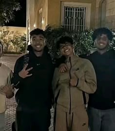 three men standing next to each other in front of a building at night with their arms around one another