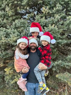 a man and two children wearing christmas hats standing in front of pine trees with their arms around each other