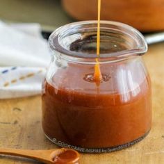 a spoon with some sauce in it on a wooden table next to a jar filled with something