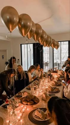 a group of people sitting around a long table with plates and candles on it, surrounded by balloons