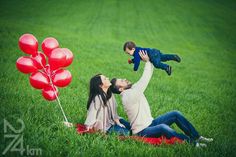 a man and woman are sitting in the grass with red balloons on their heads as a baby sits on his mother's shoulders