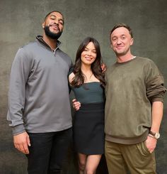 two men and a woman standing next to each other in front of a gray wall