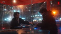 two people sitting at a table in front of a glass wall with lights on it