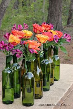 several green glass bottles with flowers in them on a wooden table near some trees and grass