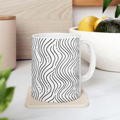 a white coffee mug sitting on top of a counter next to a bowl of fruit