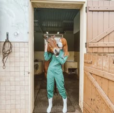 a woman in green jumpsuit standing next to a horse inside an open barn door