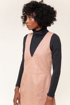 a woman with curly hair wearing a pink dress and black turtle - neck sweater, standing in front of a white background