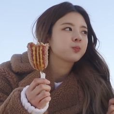 a woman in a brown coat eating a hot dog and holding a lollipop