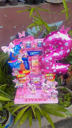 a pink birthday cake with lots of candy on it's stand in front of some plants