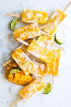 mango lassi popsicles on a white surface with lime wedges and sliced mangoes