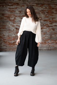 a woman standing in front of a brick wall wearing black pants and a white sweater