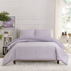 a bed with purple comforter and pillows in a white room next to a window