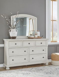 a white dresser sitting next to a window with a vase and mirror on top of it