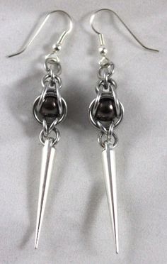 This product of Cassie's Creations is made from silver anodized aluminum rings that cage in a brown pearl bead, with silver spike charms that dangle down. The earrings are hung on black earring hooks. To get a list of the shows where we will be, visit www.MoonDragonDesigns.com, or message for custom orders Caged Crystal Earrings, Chain Maille Patterns Jewelry Making Tools, Chain Maille Patterns Earrings, Chain Mail Earrings Lone Rock Jewelry, Chainmail Patterns Earrings, Beaded Earrings Ideas, Chainmaille Jewelry Patterns, Black Earring, Jump Ring Jewelry