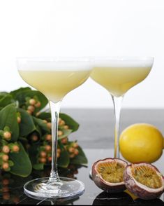 two glasses filled with yellow liquid next to lemons on a black counter top and greenery in the background