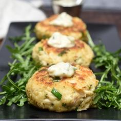 three crab cakes on a black plate topped with greens and sour cream sauce, ready to be eaten